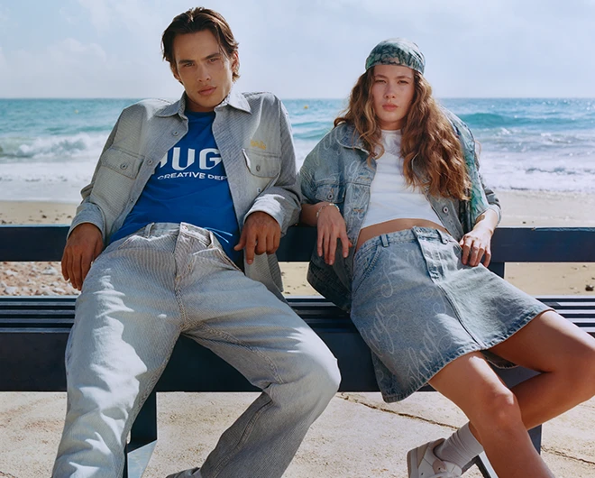 Ein junger Mann und eine junge Frau auf einer Bank am Strand in Jeans-Outfits (Foto)